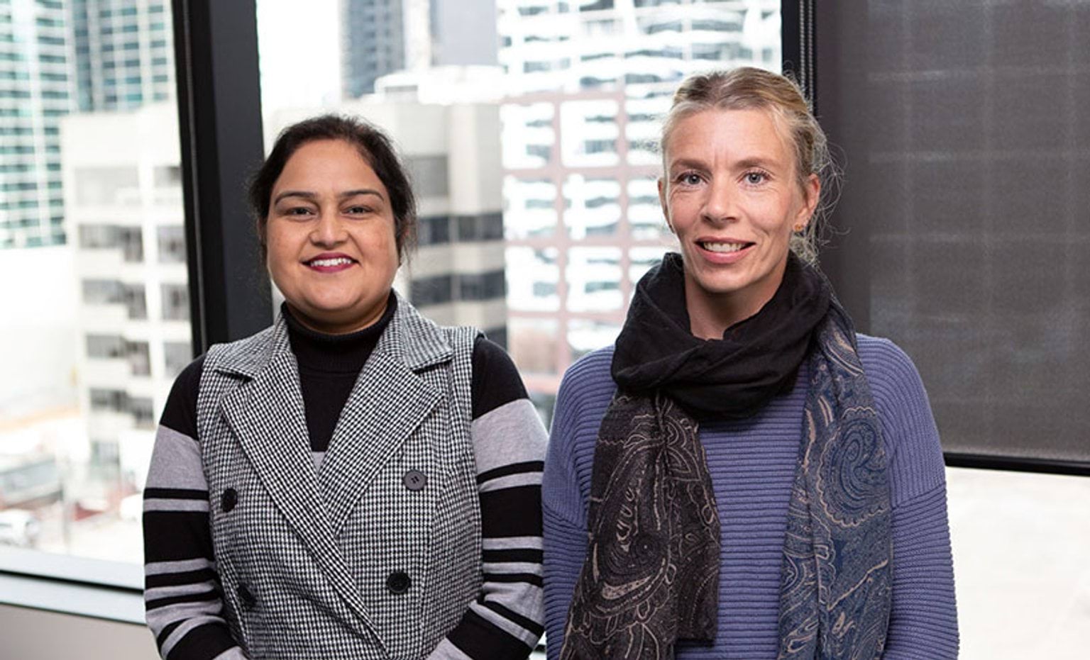 Dr Ruchita and Karla Randle standing in front of 10th floor office window with office buildings in background.