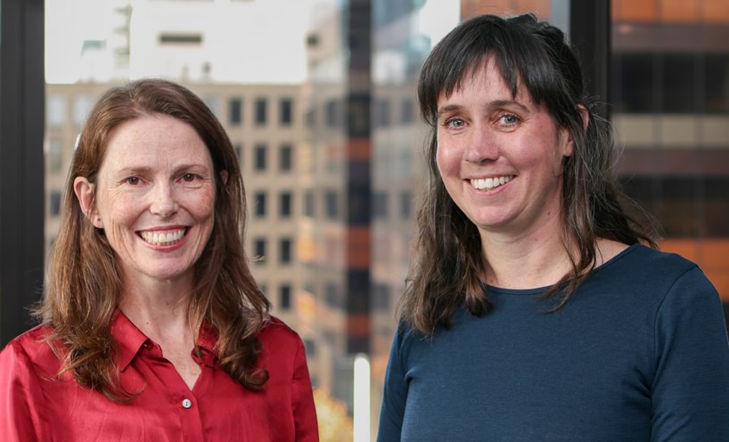Two women are smiling towards the camera. Elanor, on the left, has a deep red shirt, long auburn hair, brown eyes and pale skin with freckles. Jen, on the right, has a dark blue top, black hair with a fringe, blue eyes and pale skin.