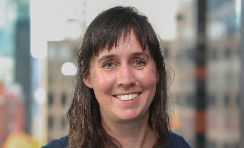 Jen is smiling broadly towards the camera. She has blue eyes and dark shoulder-length hair and a fringe. She's wearing a dark blue top.