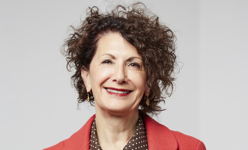 Christine has short, curly brown hair, brown eyes and fair skin. She's smiling widely towards the camera and wearing a dark spotted shirt and red blazer. 